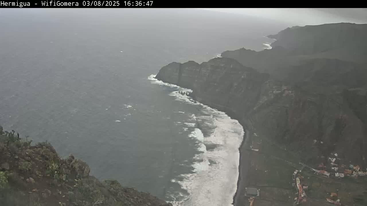 A high-angle, long shot shows a rocky coastline with crashing waves on a cloudy day.