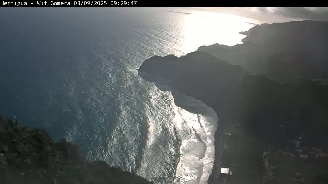 A high-angle view shows a rocky coastline meeting a bright, wavy ocean under a mostly sunny sky.