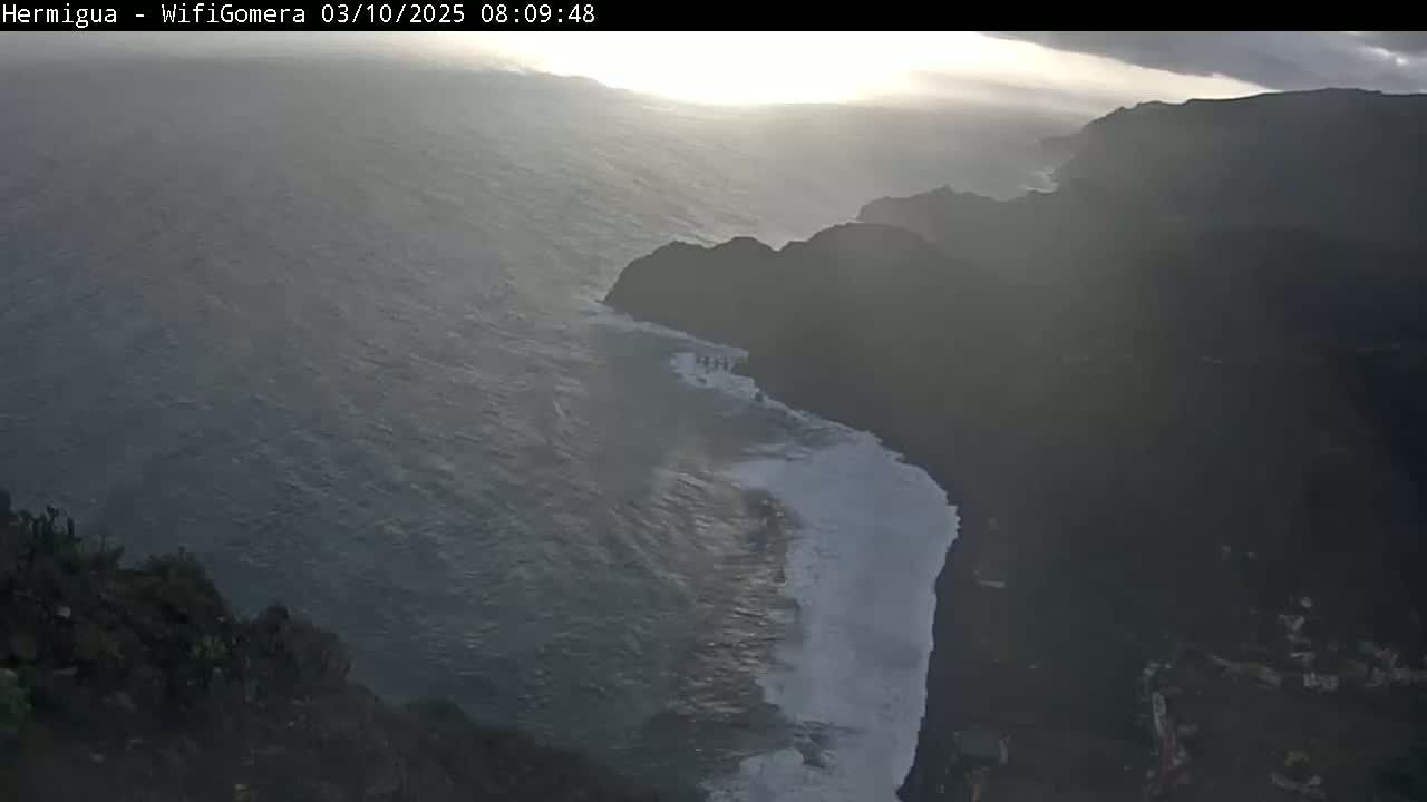 A high-angle, long shot shows a dark, rocky coastline meeting a choppy ocean under an overcast sky with a hint of sunlight breaking through the clouds.