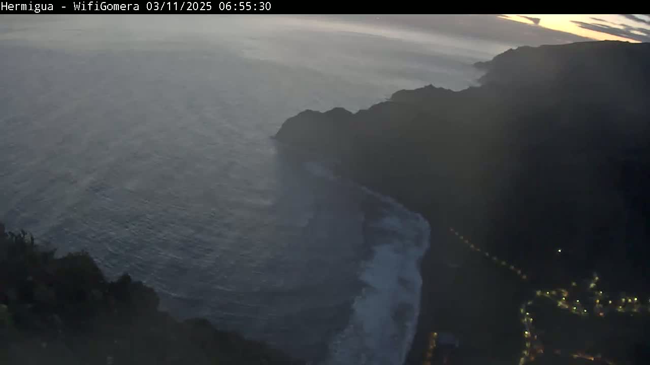 A high-angle, twilight view shows a dark, rocky coastline with waves crashing against it, overlooking a relatively calm ocean.