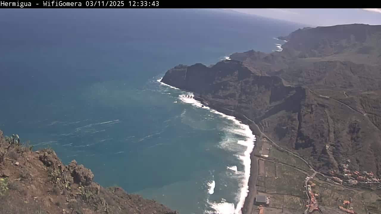 A high-angle view shows a rocky coastline meeting a dark-blue ocean with white-capped waves under a clear, sunny sky.