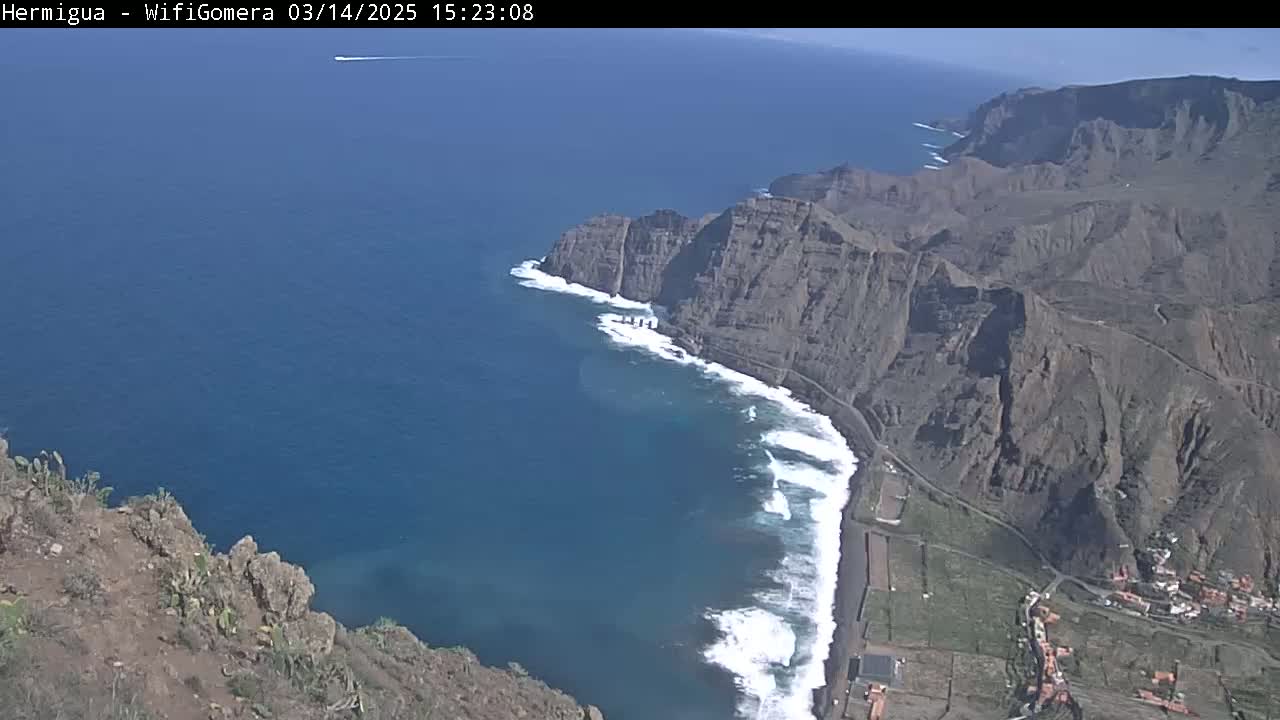 A high-angle view shows a rocky, dark-brown coastline meeting a deep-blue ocean with white-capped waves under a clear, sunny sky.