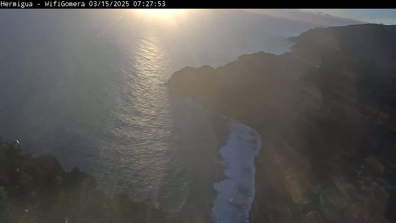 A hazy sunrise illuminates a dark rocky coastline meeting a calm, sunlit ocean.