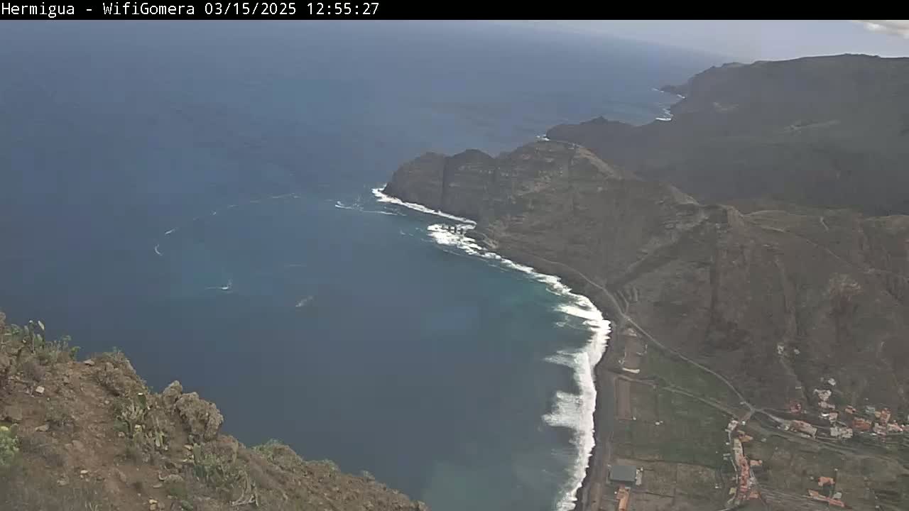 A high-angle view shows dark-colored, rocky cliffs meeting a dark-blue ocean with white waves along the shore, under somewhat hazy skies.