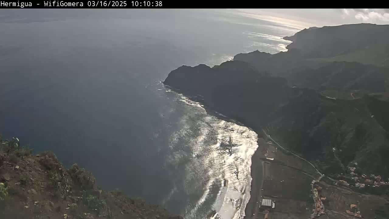 A high-angle view shows a dramatic coastline with dark volcanic cliffs meeting a sea with waves breaking on a beach under a mostly clear sky.