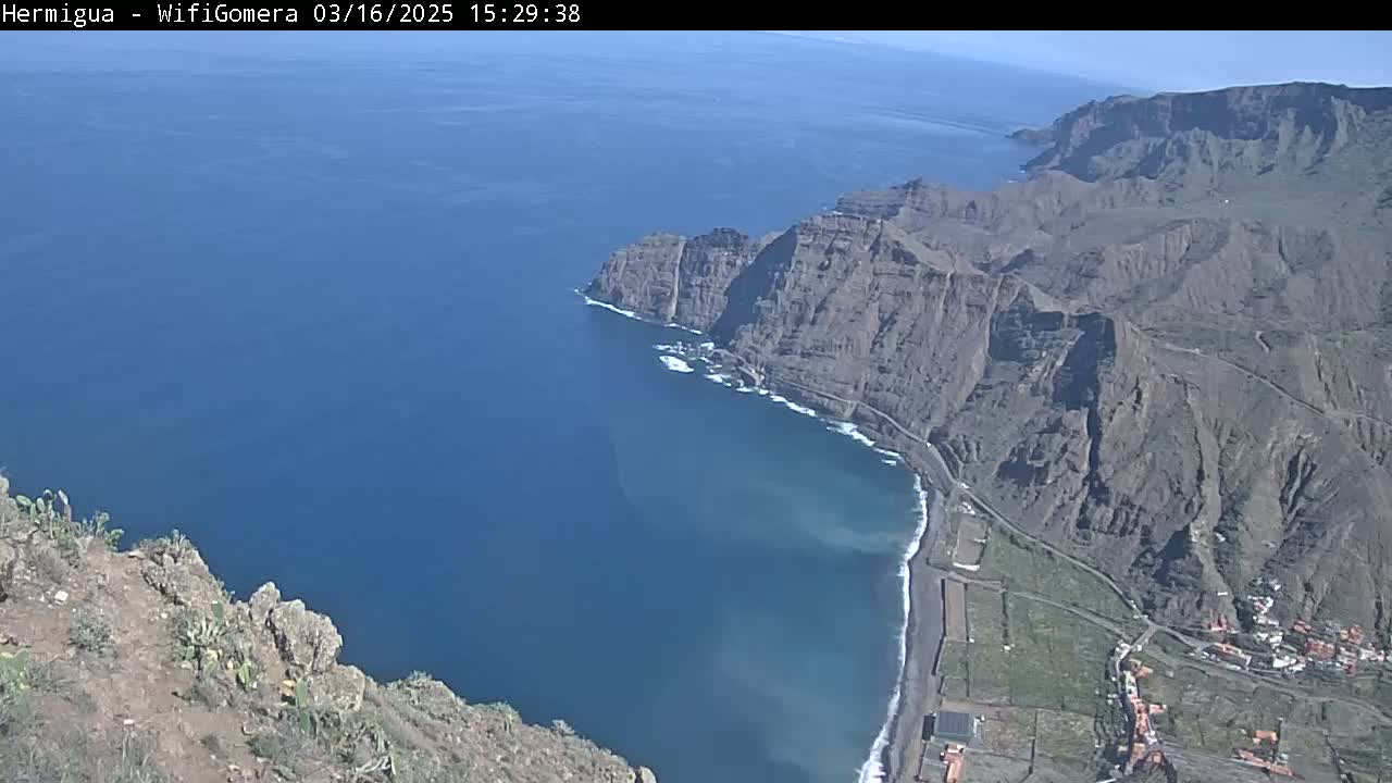 A high-angle view on a sunny day shows a dark-colored, rocky coastline meeting a deep blue ocean; a small beach is visible along the coast, with sparse vegetation inland.