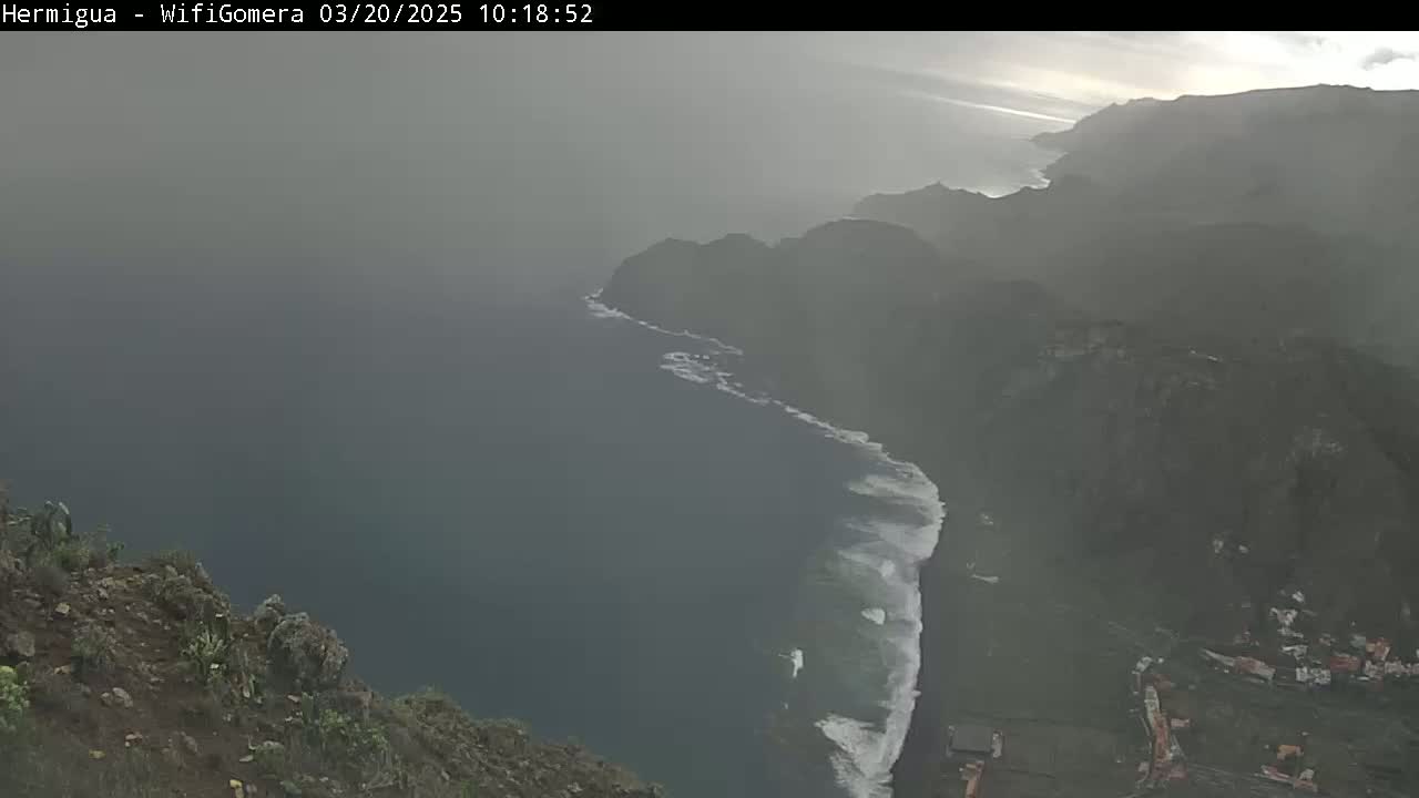 A hazy, overcast view shows a rocky coastline with waves crashing on a dark beach, seen from a high vantage point.