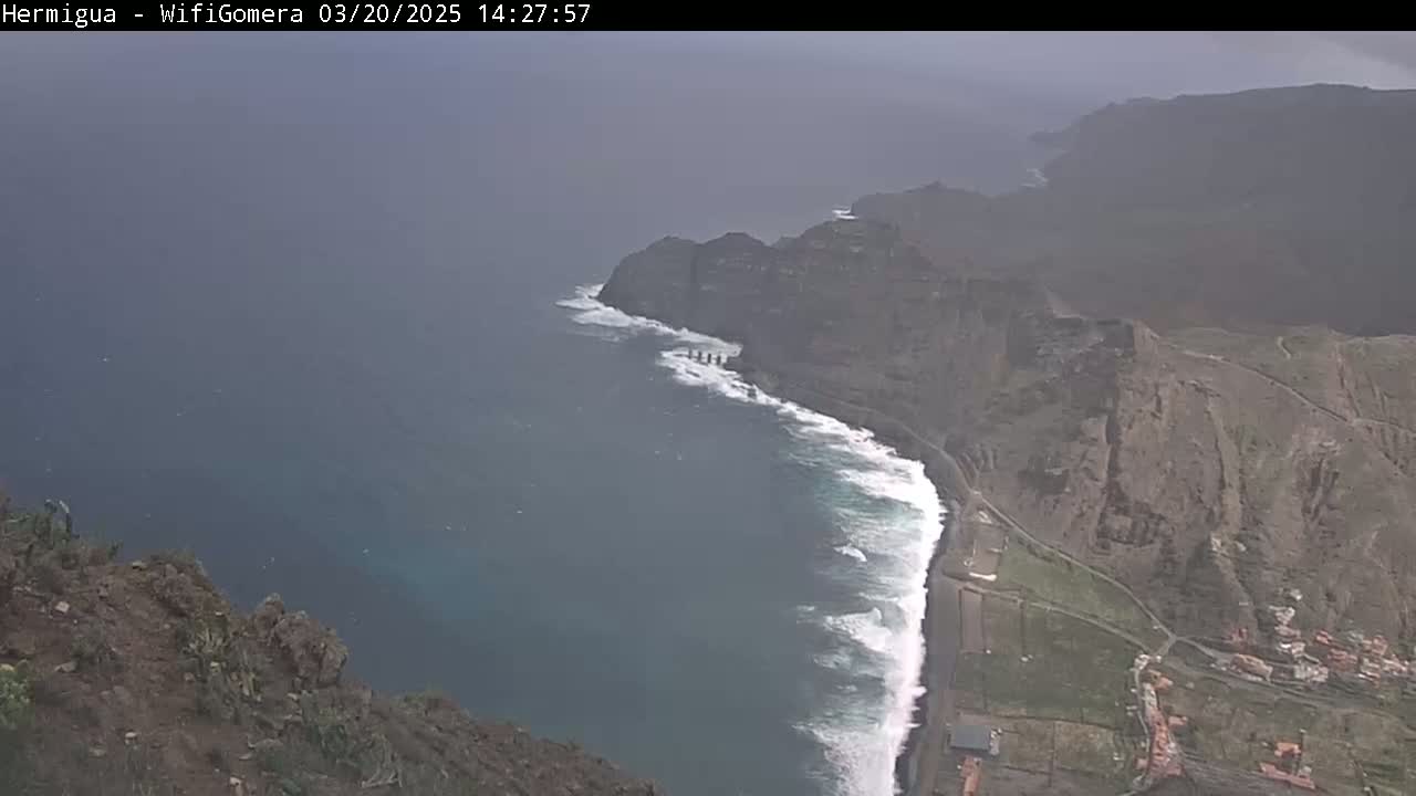 A hazy, overcast day reveals a rocky coastline with crashing waves along a dark-blue ocean, and a village nestled against the cliffs.