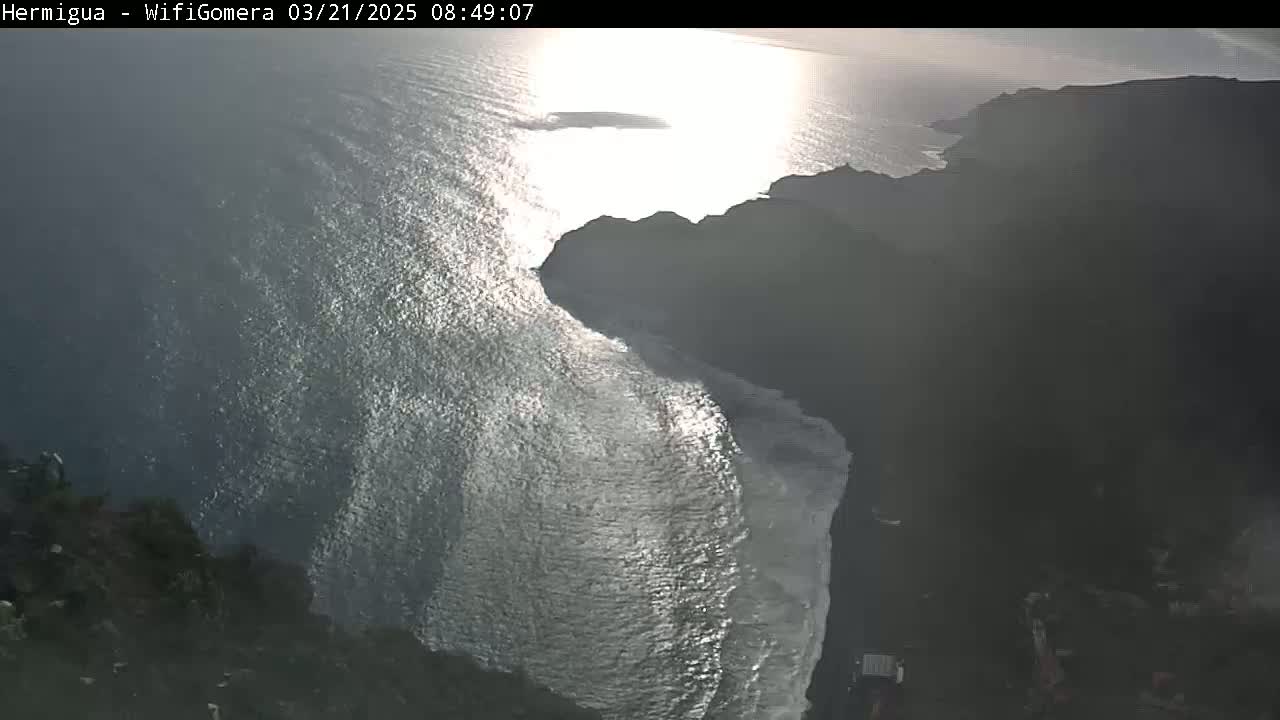 A bright, sunny day shows a view of dark, rocky cliffs meeting a sparkling ocean, with waves breaking against the shore.