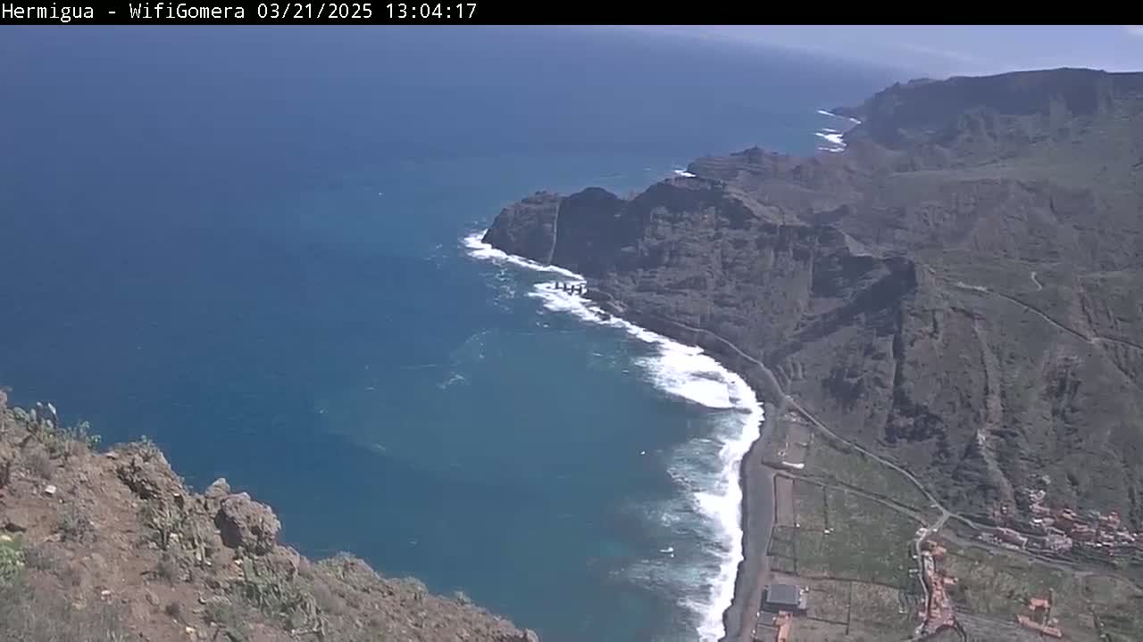 A high-angle view shows a dark-colored, rocky coastline meeting a blue ocean with white waves under a mostly sunny sky.