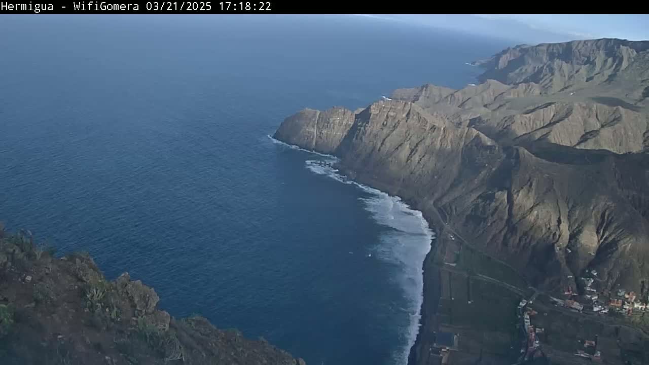 A high-angle view shows a dramatic coastline with dark, rocky cliffs meeting a dark blue ocean where white waves are crashing onto a beach.