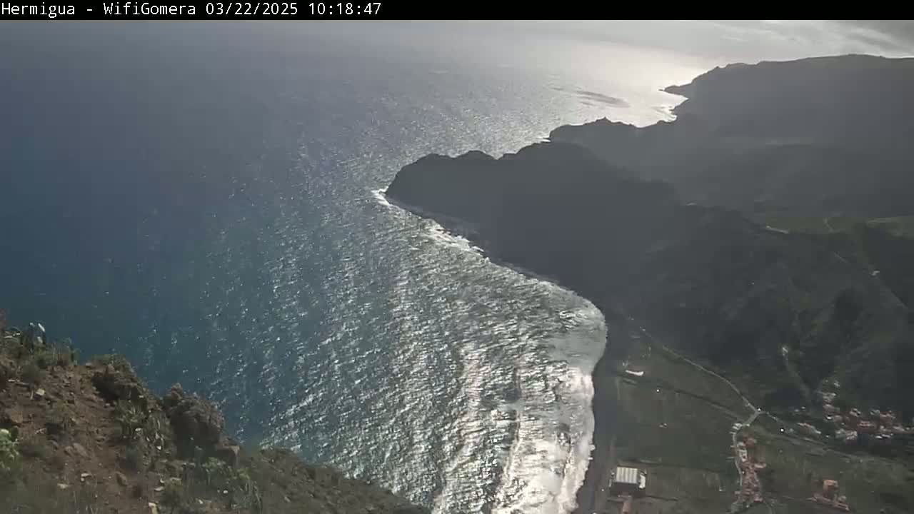 A high-angle view shows a dark, rocky coastline meeting a bright blue ocean with waves crashing on the shore under a mostly sunny sky.