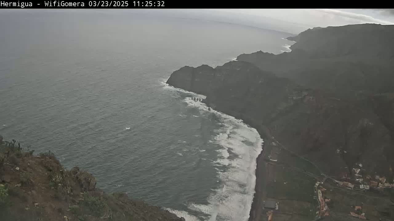 A high-angle, long shot shows a dark-colored, rocky coastline with crashing waves on an overcast day.