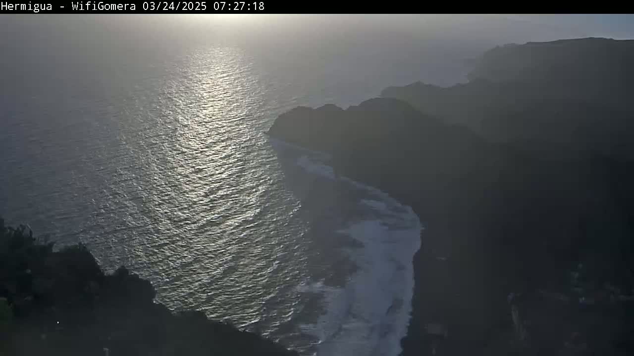 A high-angle view shows a dark, rocky coastline meeting a sunlit ocean, with hazy weather conditions.