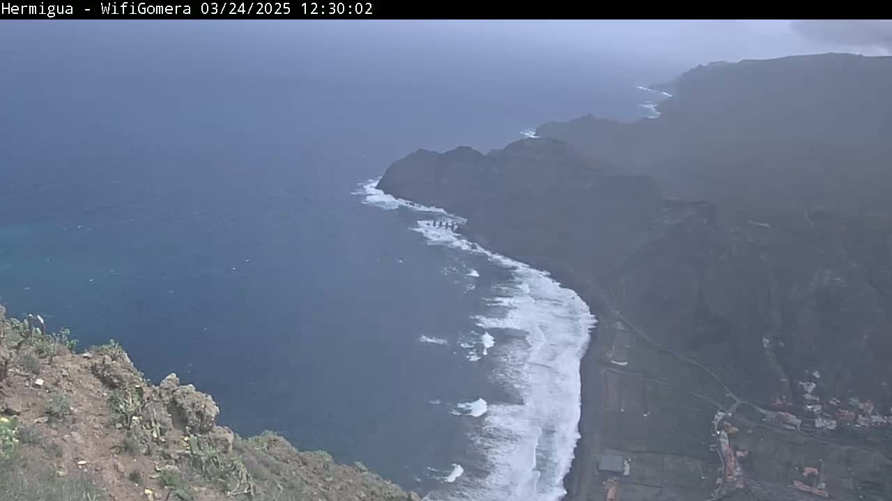 A hazy coastal view shows dark, rocky cliffs meeting a dark-blue ocean where white waves crash against a dark-sand beach.