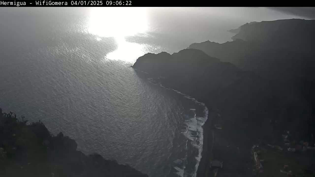 A hazy, sunlit ocean meets dark, rocky cliffs under overcast skies.