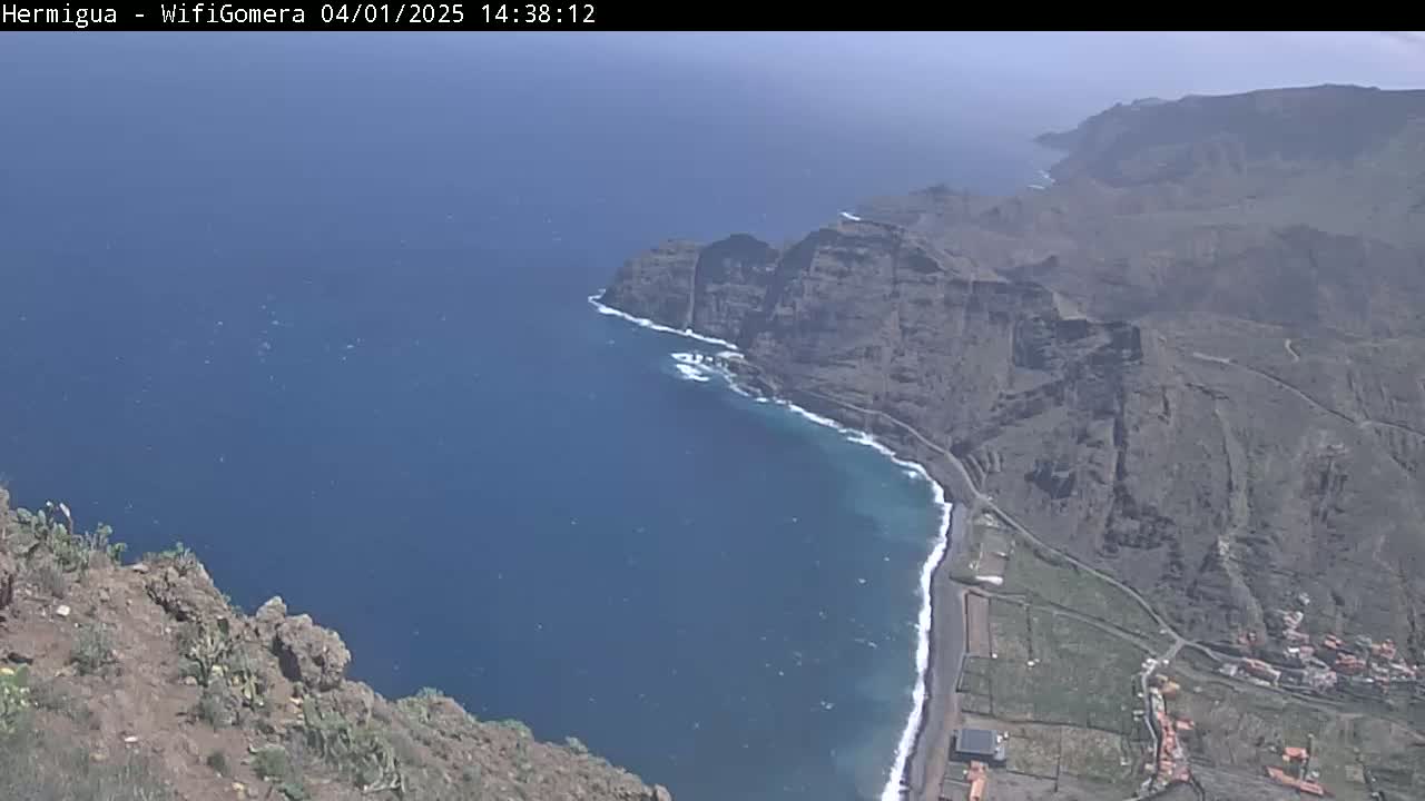 A high-angle view shows a dark-blue ocean meeting a dark-brown, rocky coastline under a mostly clear sky.