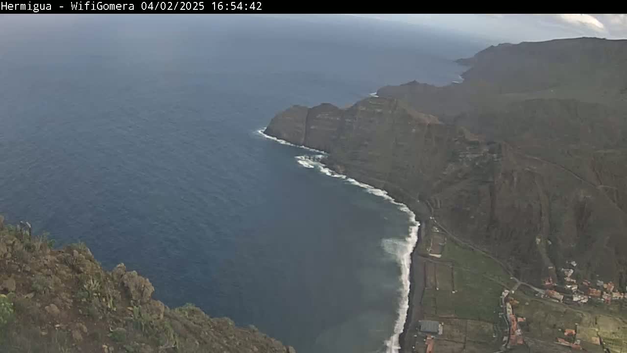 A high-angle view shows dark-colored cliffs meeting a dark-blue ocean with small waves, and a sliver of sandy beach, under a mostly cloudy sky.