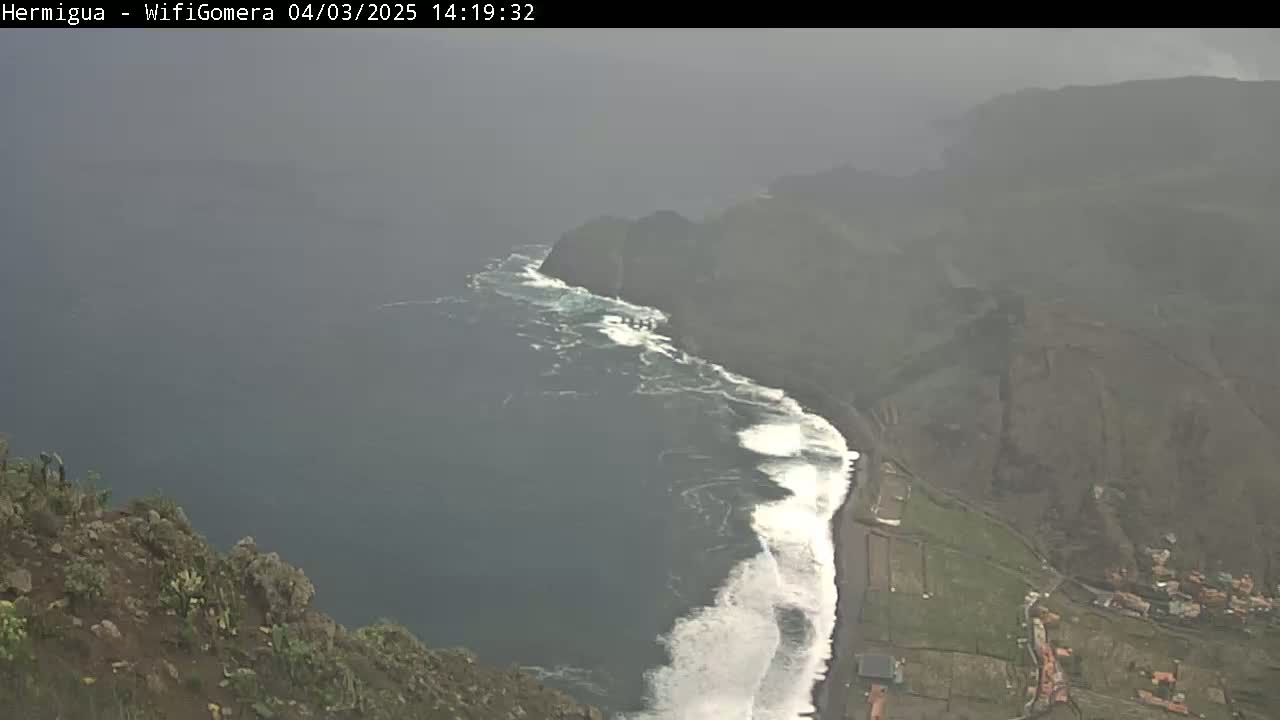 A hazy, overcast view shows a rocky coastline with waves crashing on a dark-sand beach below a cliff, where some small buildings are visible.