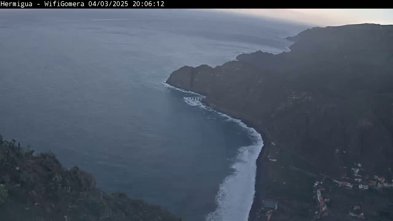 A high-angle, twilight view shows a dark, rocky coastline meeting a dark-blue ocean with white waves crashing against the shore.