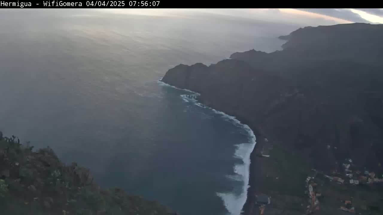 A hazy sunrise illuminates a dark volcanic coastline where waves crash against a black sand beach.