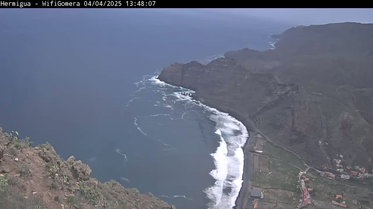 A high-angle view shows a dark, rocky coastline with a dark-sand beach where waves are crashing, all under an overcast sky.