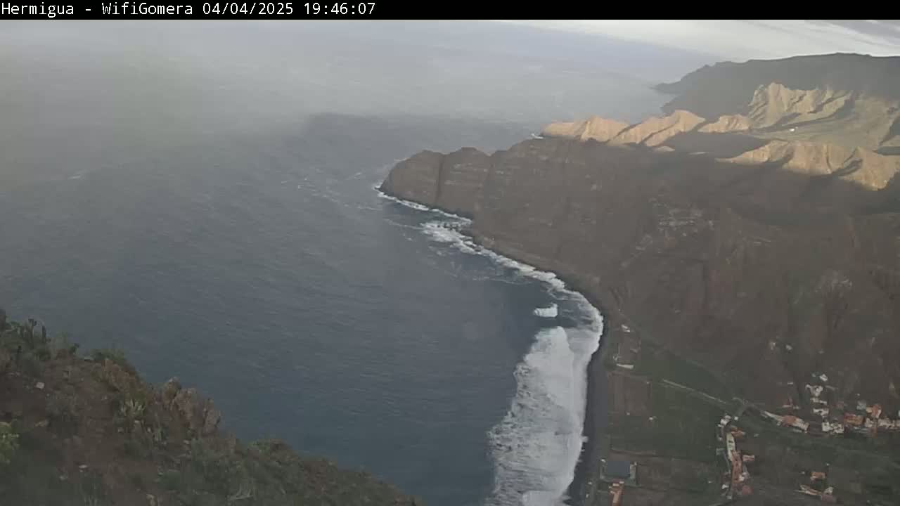 A hazy coastal view shows dark, rocky cliffs meeting a dark ocean with white waves crashing along the shore.
