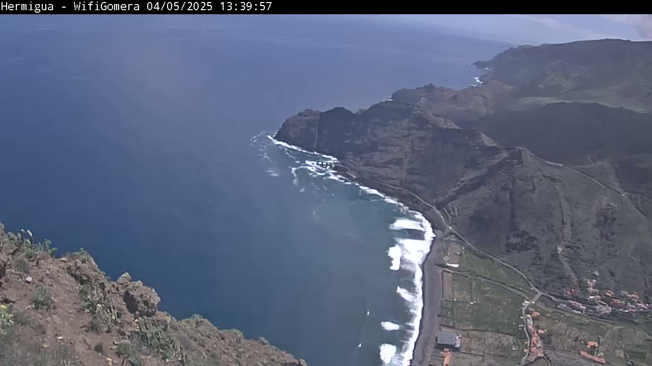 A high-angle view shows a rocky coastline with a dark-sand beach meeting a dark-blue ocean under a mostly clear sky.