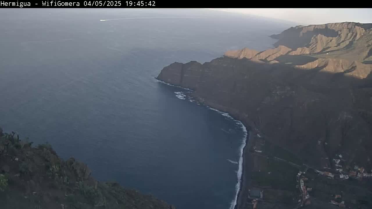 A high-angle view shows dark, rocky cliffs meeting a calm, dark-blue ocean under a mostly clear, dusky sky.