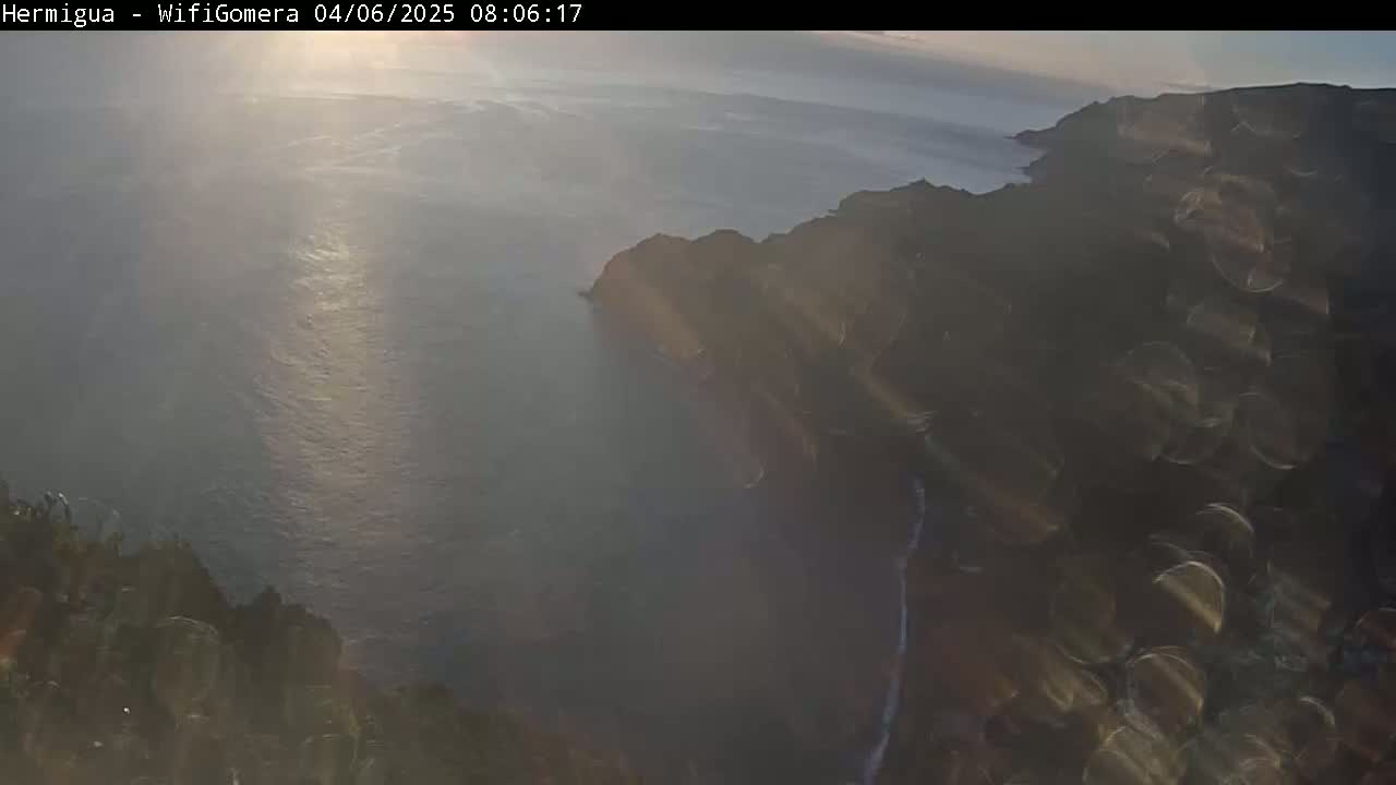 A hazy sunrise illuminates a dark, rocky coastline meeting a calm, sunlit ocean.