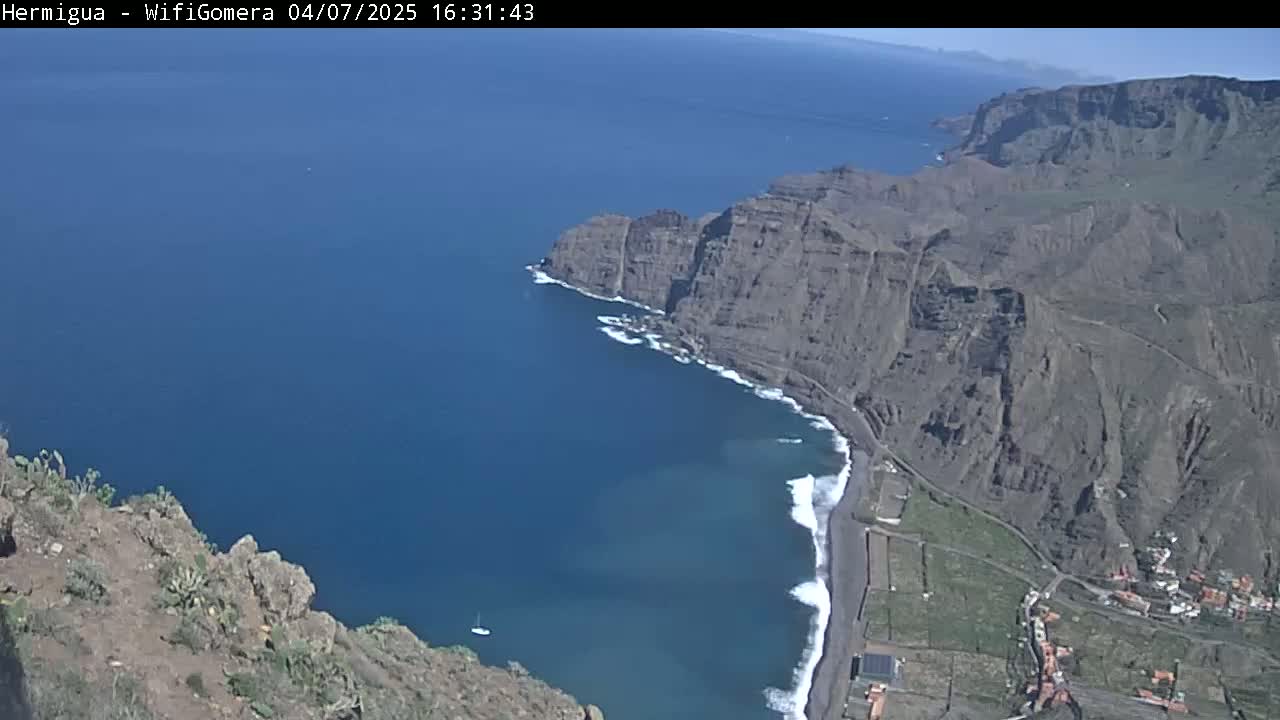 High-angle view of a dramatic coastline with dark cliffs meeting a deep blue ocean on a sunny day, featuring a small beach with white waves.