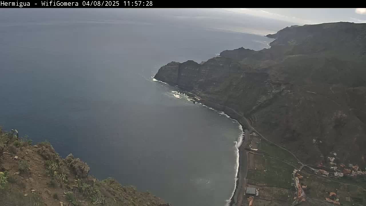 A high-angle view shows a dark-grey ocean meeting a rocky, dark-brown coastline with a small town nestled at the edge of the water under partly cloudy skies.