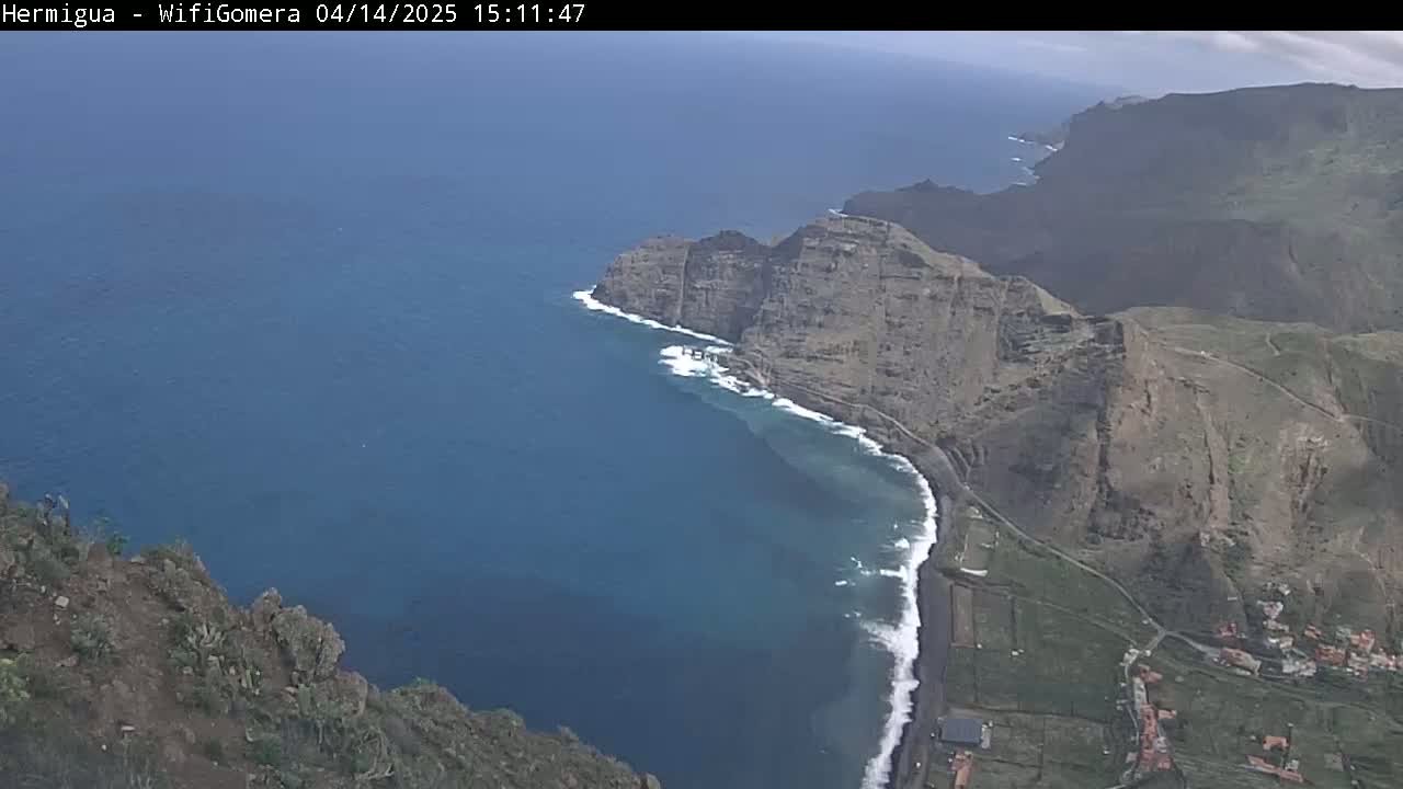 A high-angle view shows a dark-blue ocean meeting a rocky, brown coastline with small villages under a mostly clear sky.