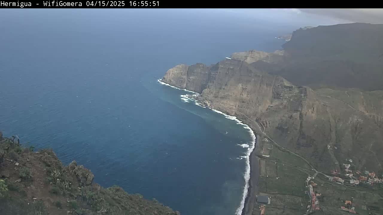 A high-angle view shows a dark-blue ocean meeting a dark-brown, rocky coastline with a small, dark beach under mostly cloudy skies.