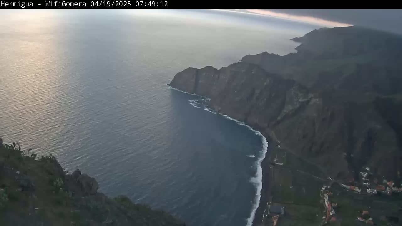 A high-angle view shows dark, rocky cliffs meeting a dark-blue ocean with small waves under an overcast sky.