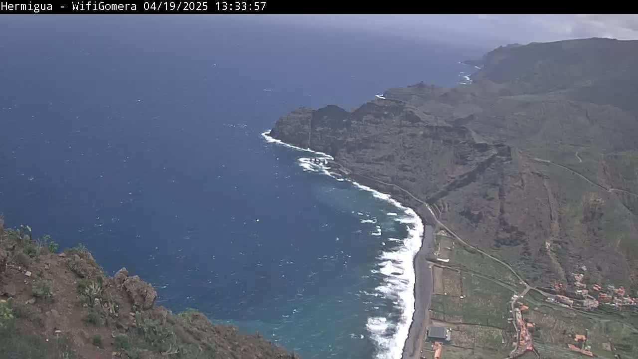 A high-angle view shows a rocky coastline meeting a dark-blue ocean with waves breaking on a dark sand beach under a partly cloudy sky.