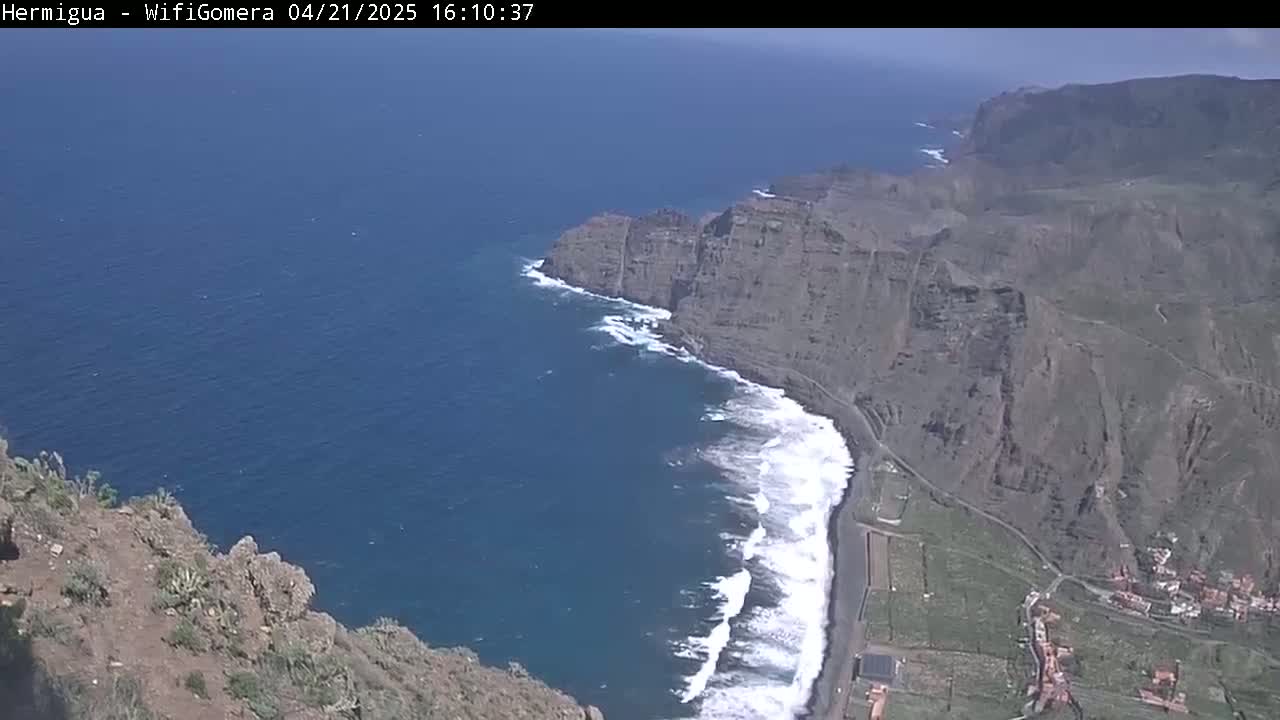 High-angle view of a rocky, dark-colored coastline meeting a deep blue ocean with white-capped waves under a clear sky.