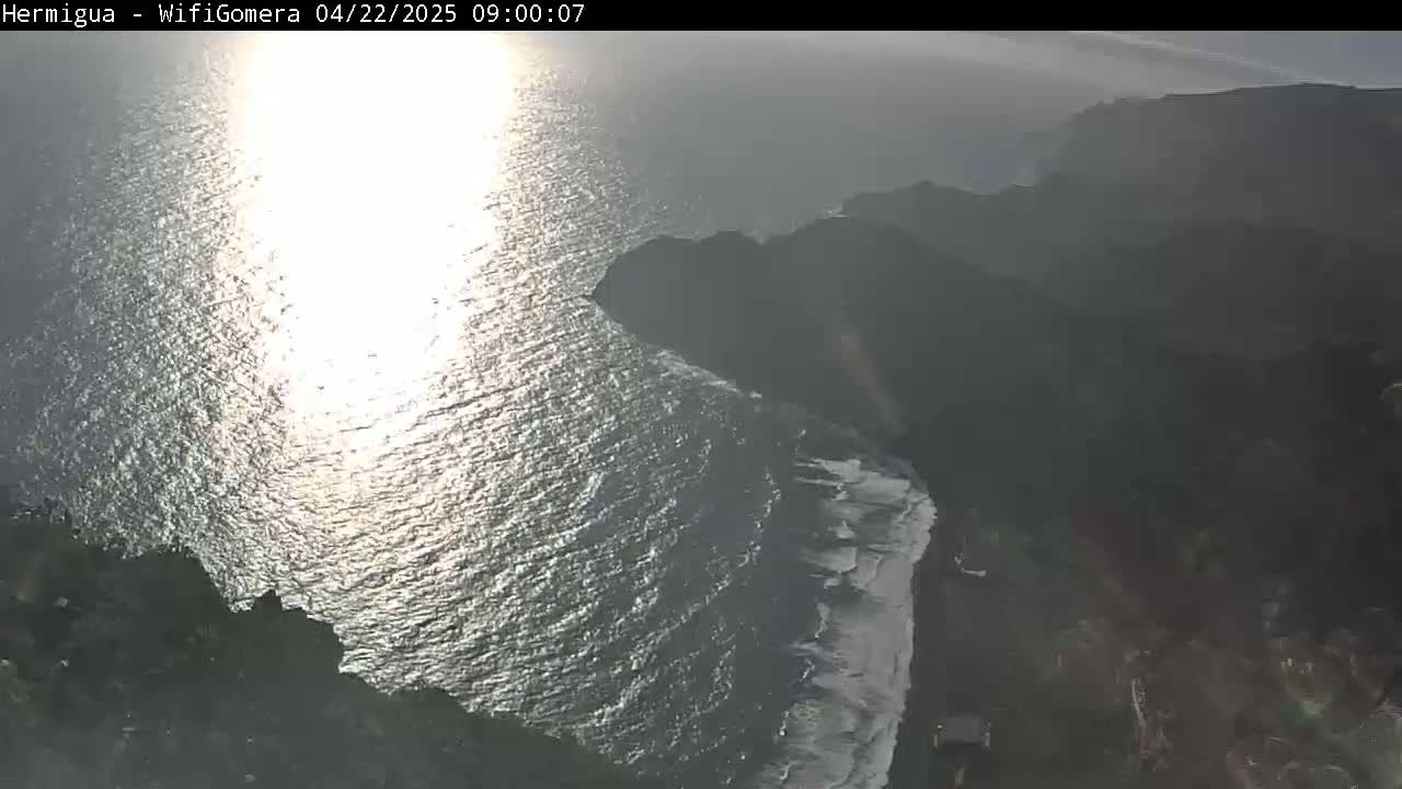A high-angle view shows a rocky coastline meeting a bright, sunlit ocean on a hazy day.
