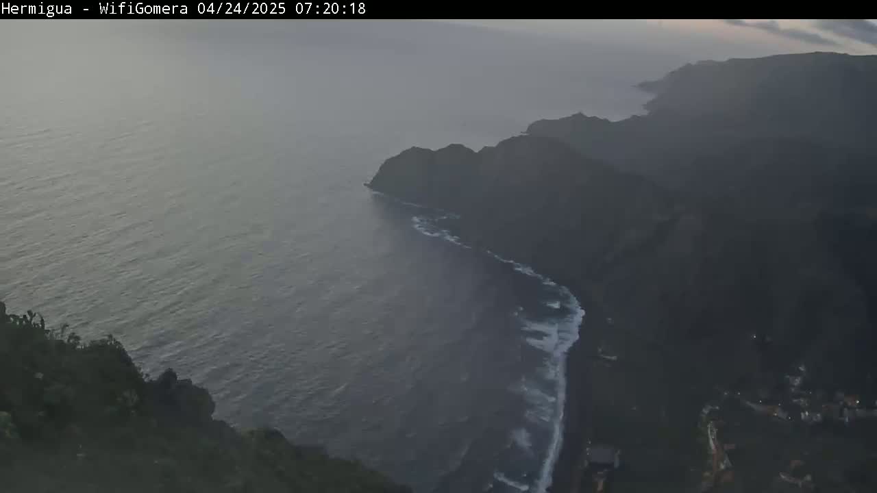 A high-angle, hazy view shows a dark, rocky coastline with waves breaking against a dark-colored beach, meeting a relatively calm, dark ocean.