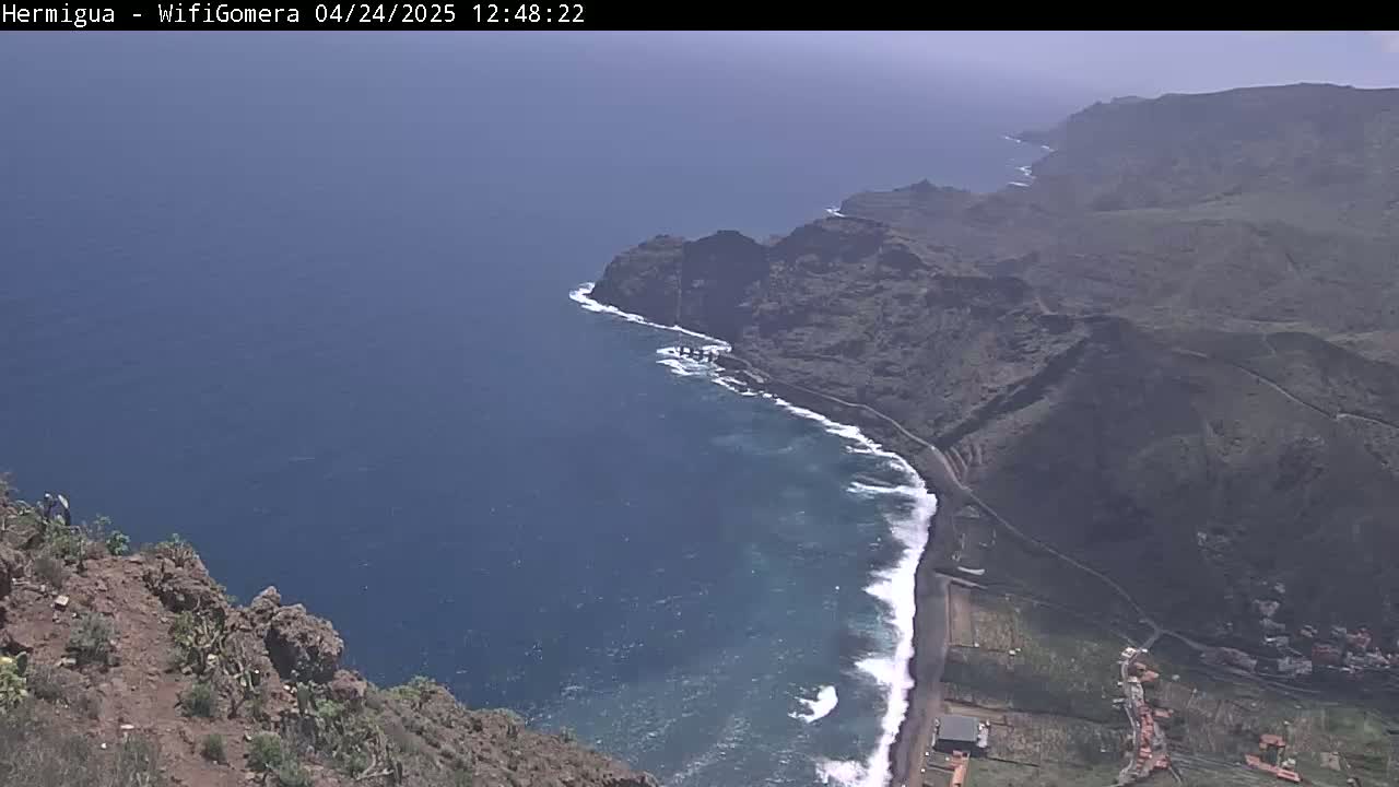 A high-angle view shows a rocky coastline meeting a dark blue ocean with white waves under a mostly clear sky.