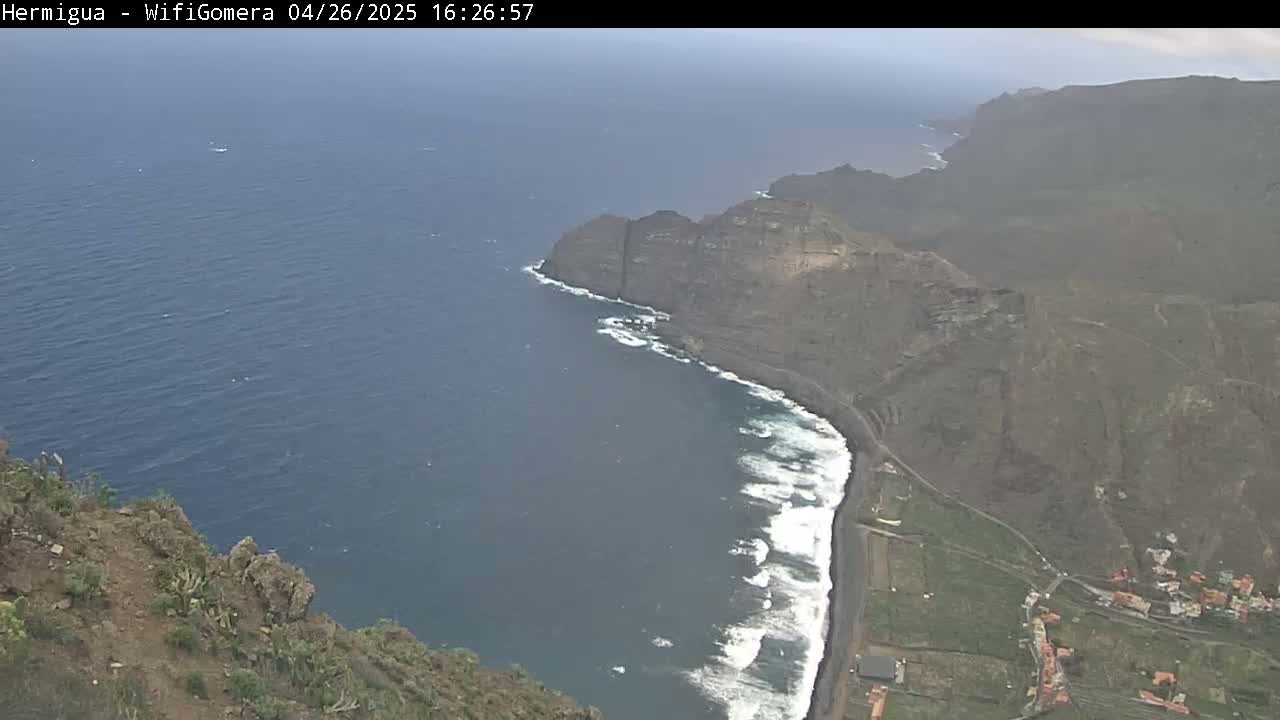 A high-angle view shows a dark-colored, rocky coastline with waves crashing on a black sand beach, under hazy conditions.