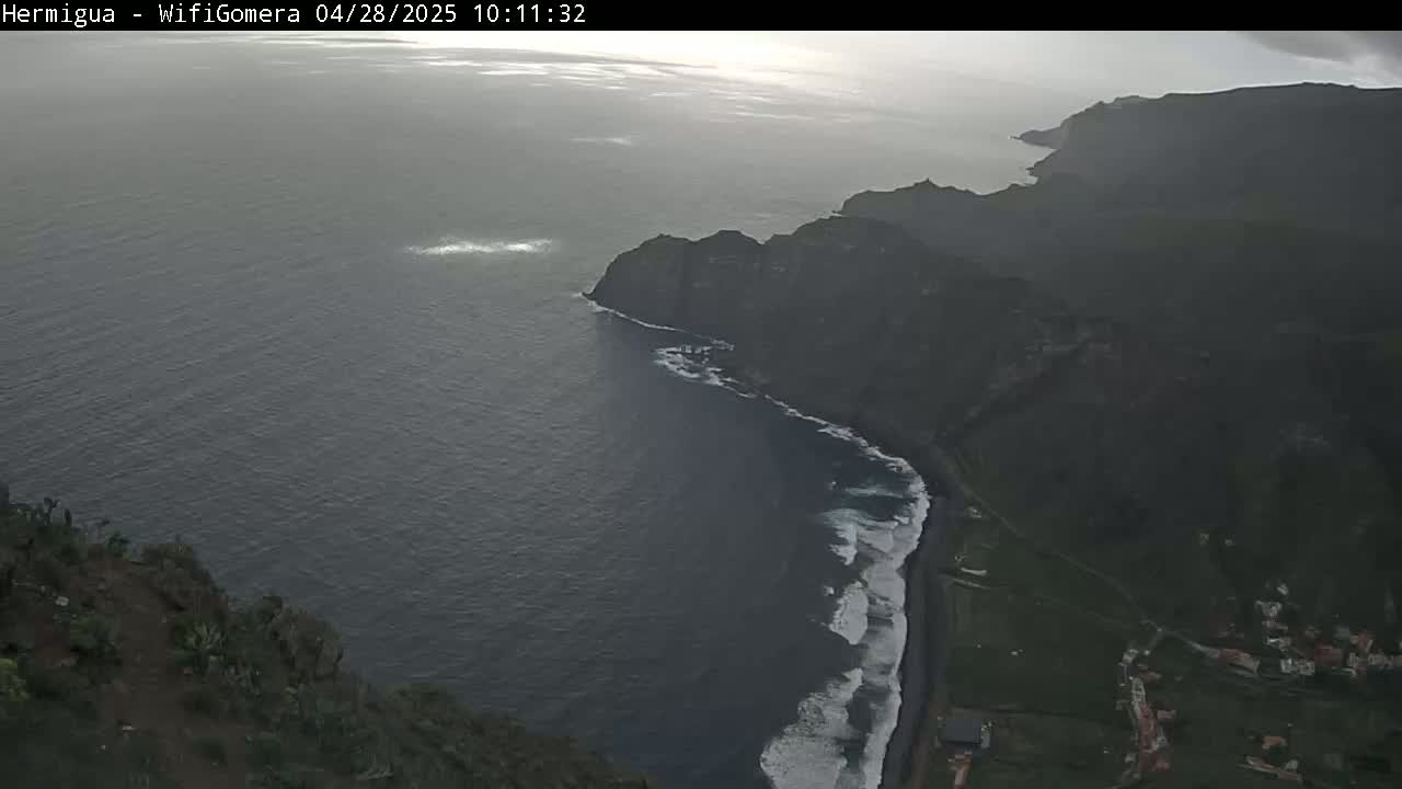 A high-angle view shows a dark, overcast day with dark-blue ocean waves lapping at a black-sand beach next to dark-green cliffs.