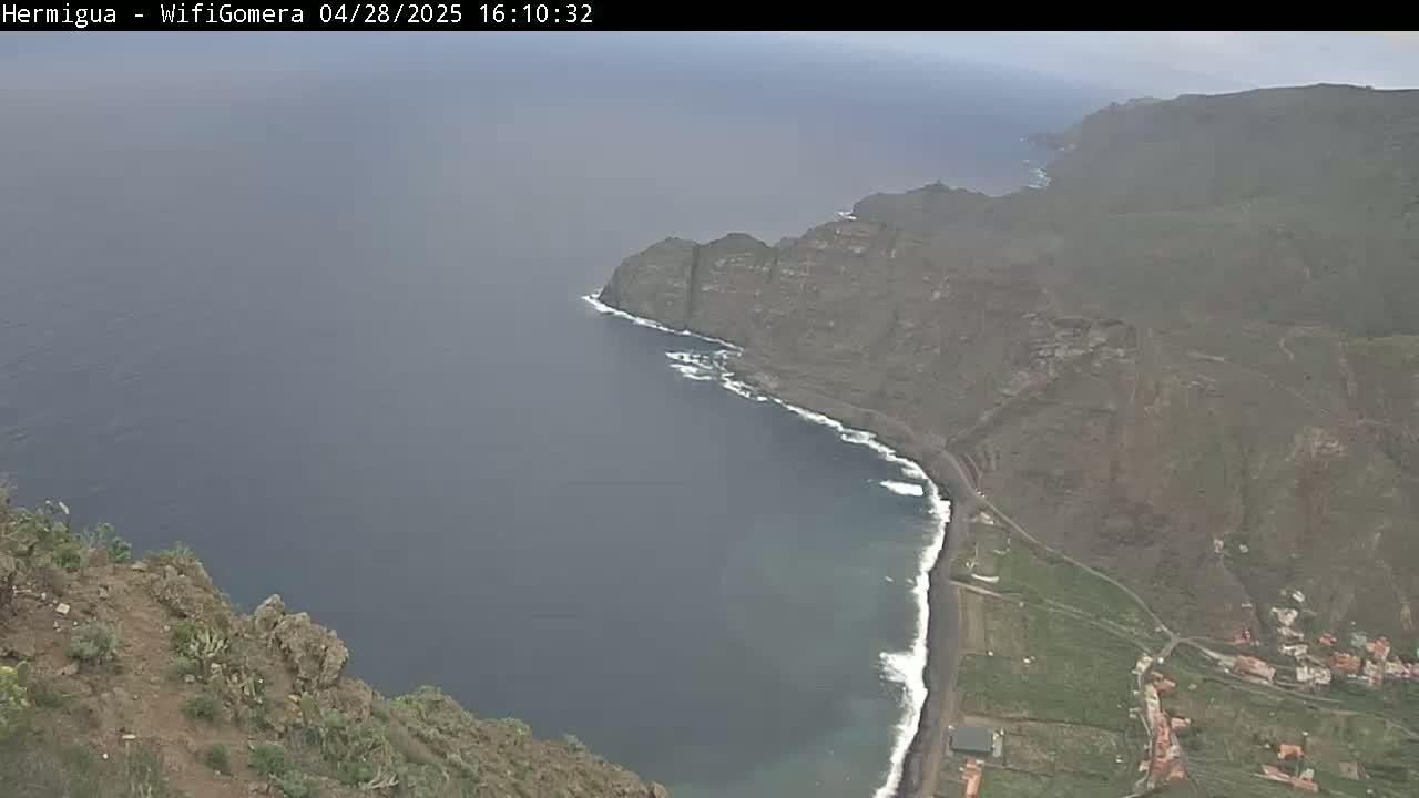 A high-angle, long shot shows a dark-grey ocean meeting a dark-brown, rocky coastline with a small village at the base of the cliffs on an overcast day.