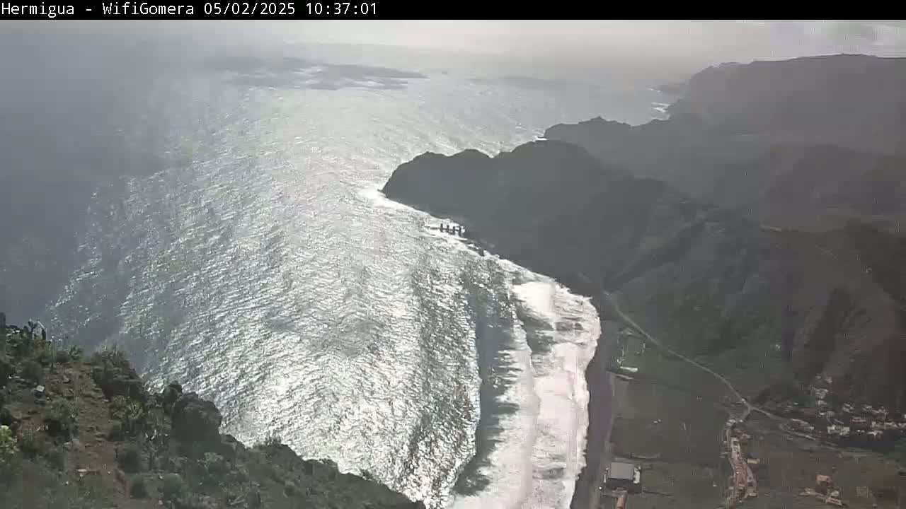 A high-angle view shows a dark, rocky coastline with a black sand beach where waves are crashing, all under an overcast sky.