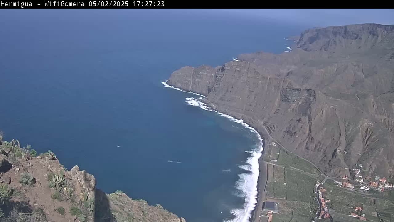 A high-angle, long shot shows a rocky, dark-colored coastline meeting a dark-blue ocean with white waves on a sunny day.