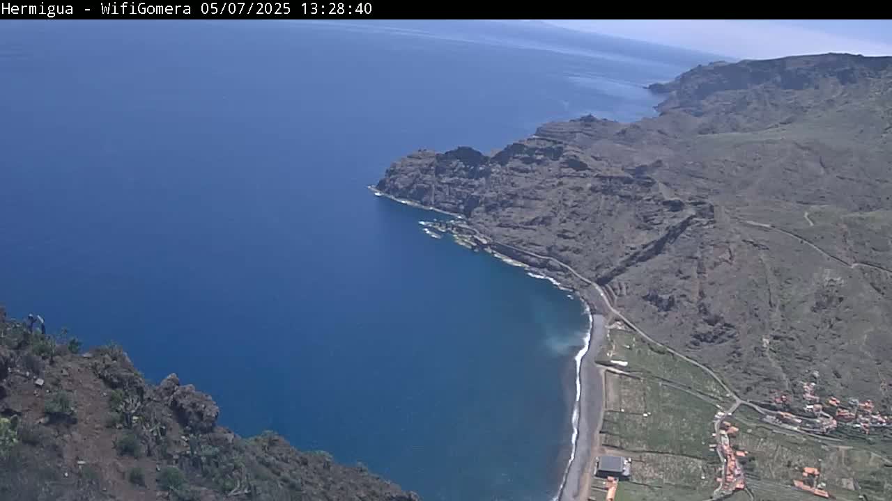 A high-angle view shows a dark-colored, rocky coastline meeting a deep blue ocean under sunny conditions.