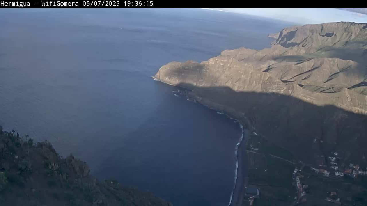 A high-angle view shows a rocky coastline meeting a calm, deep-blue ocean under a clear sky.