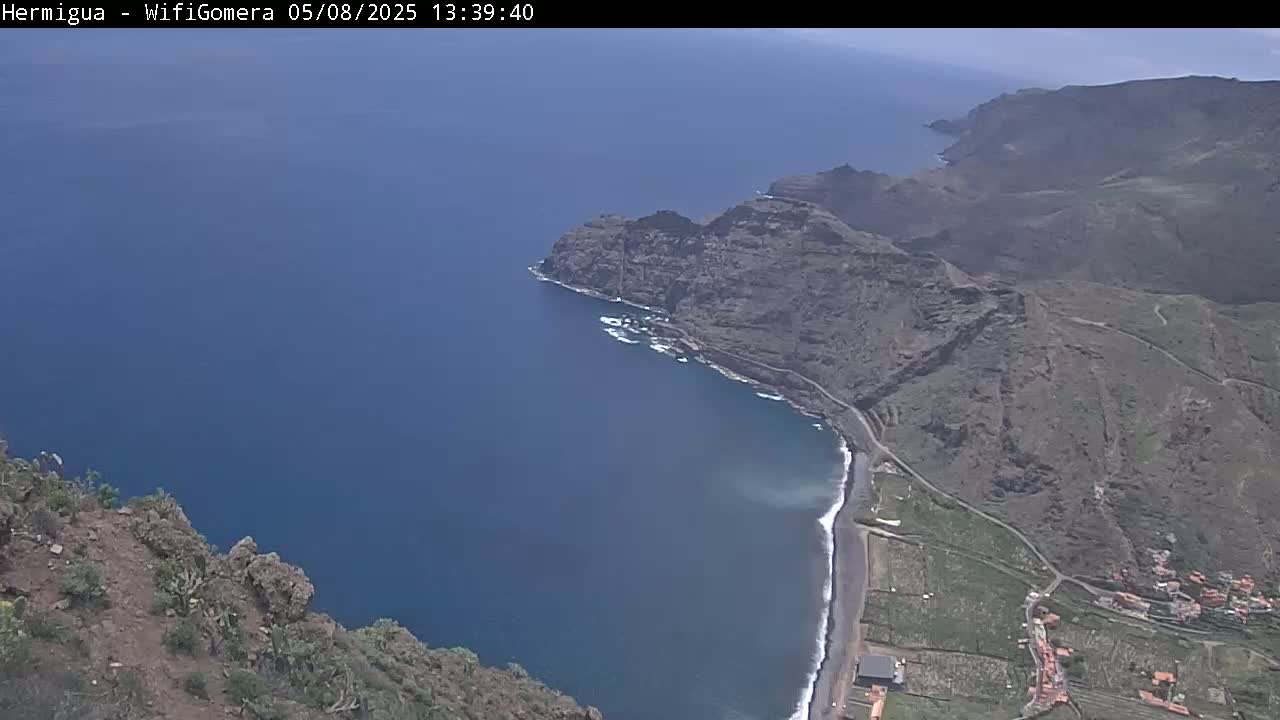 A high-angle view shows a dark-blue ocean meeting a rocky, brown coastline on a sunny day.