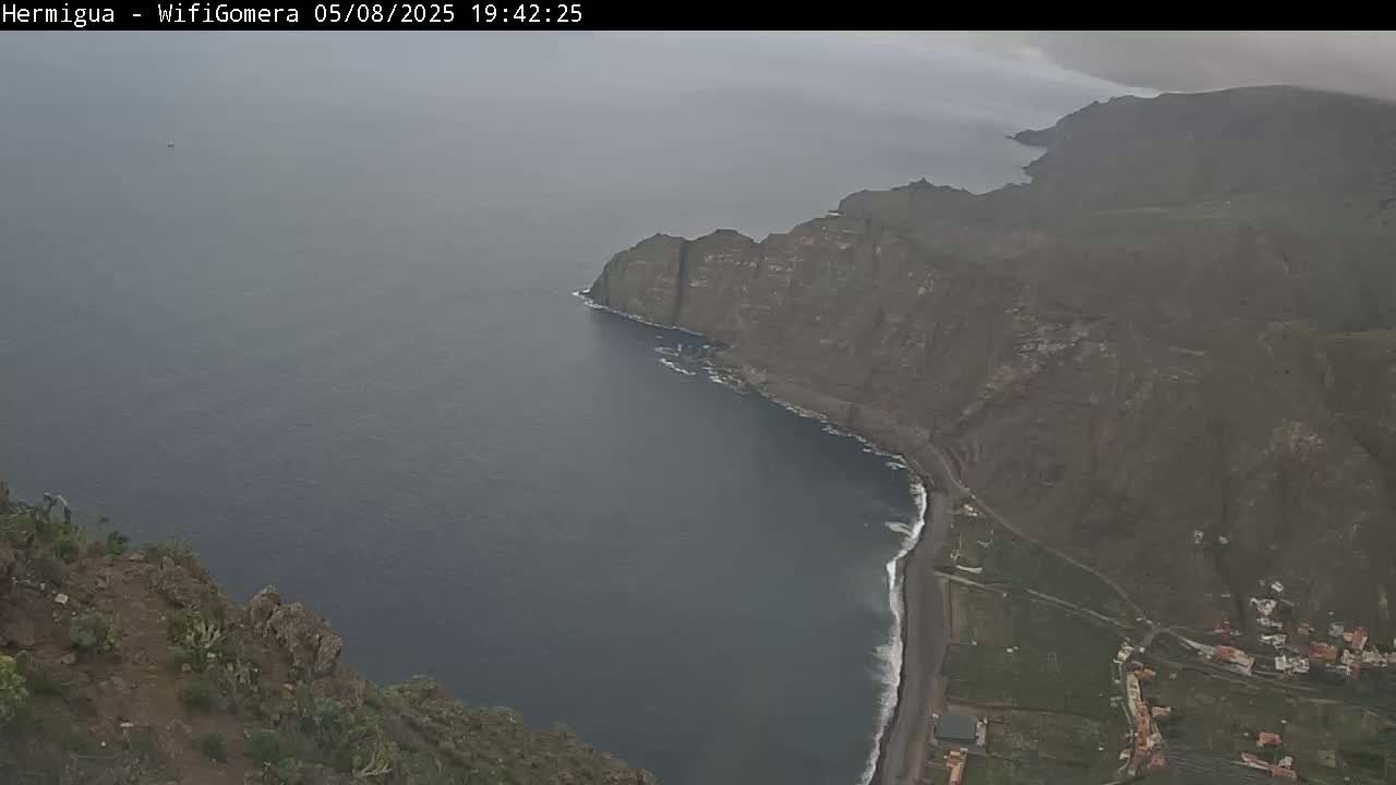 A high-angle view shows a dark-blue ocean meeting a rocky, dark-green coastline, with a small town visible on the land under an overcast sky.