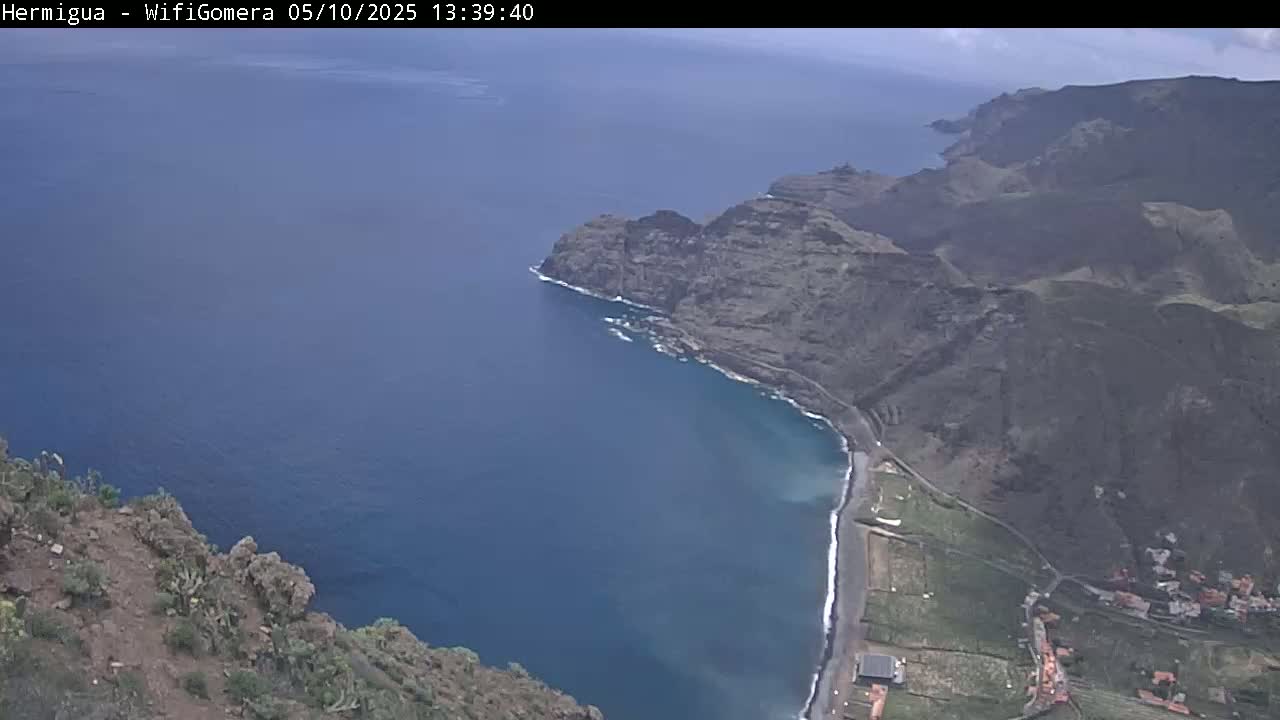 A high-angle view shows a mostly sunny day overlooking a dark-blue ocean meeting a rocky, dark-brown coastline with a small beach and some buildings.